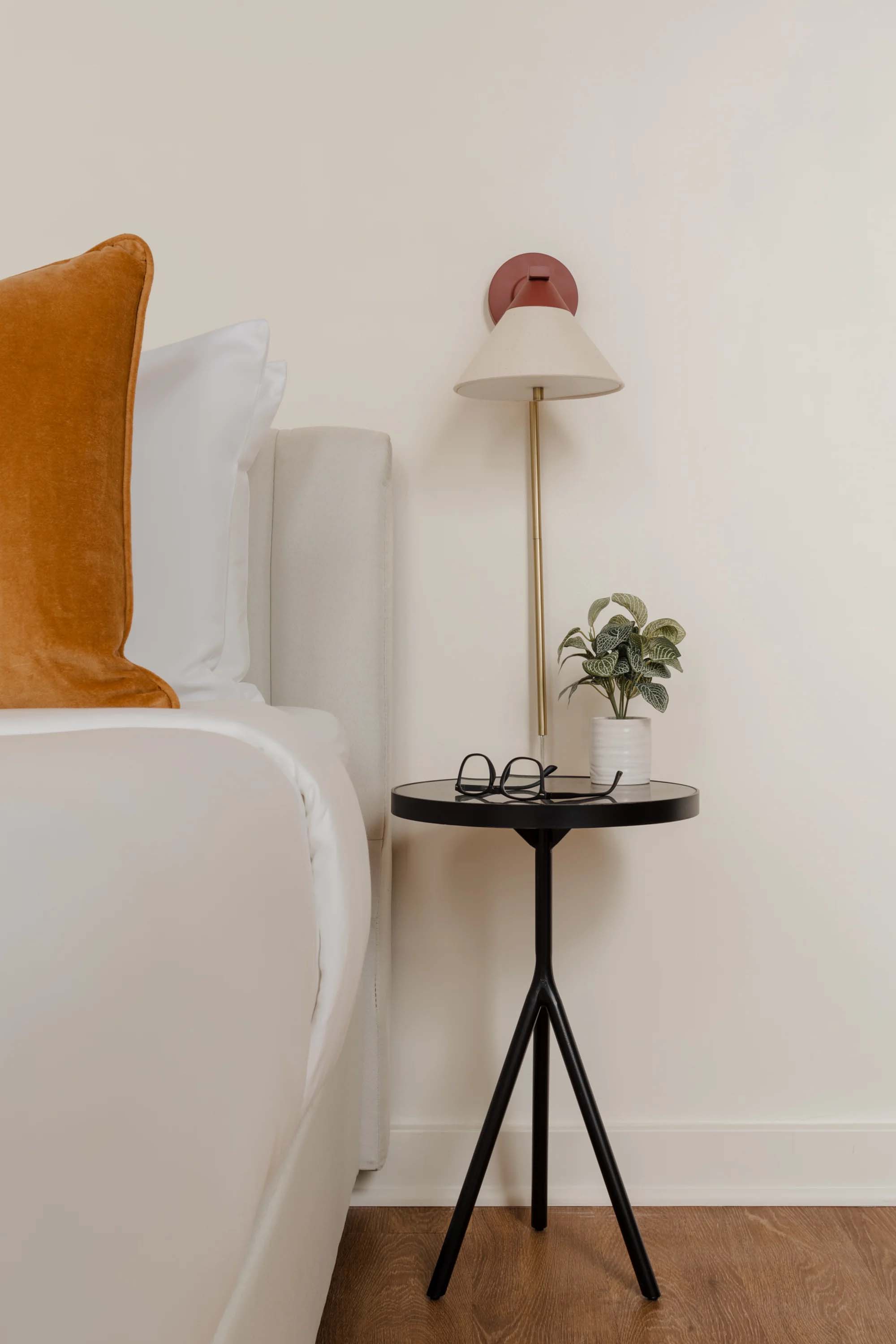 A bedside table with a lamp, plant, and glasses in a white room.